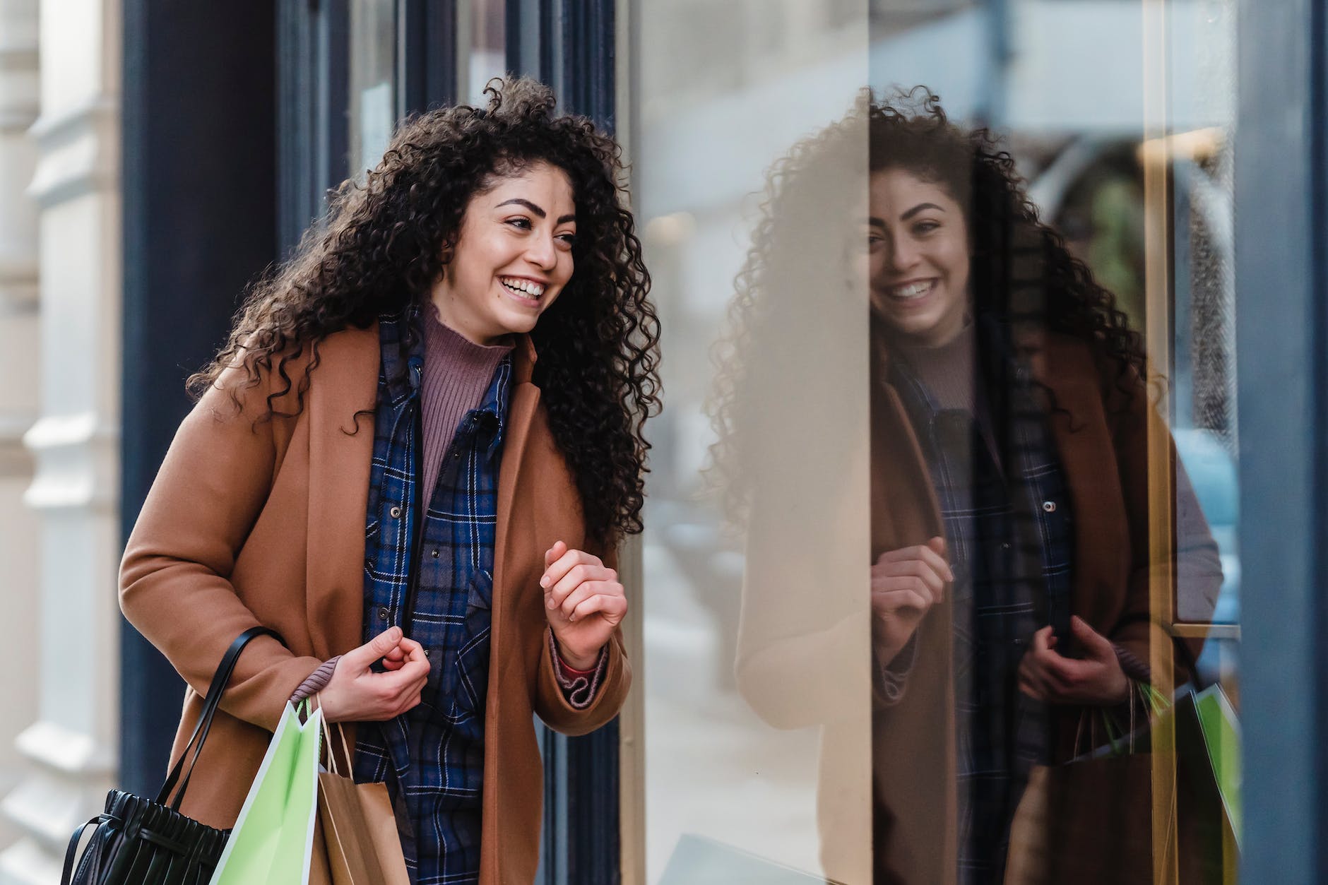 joyful ethnic woman with shopping bags standing near glass building